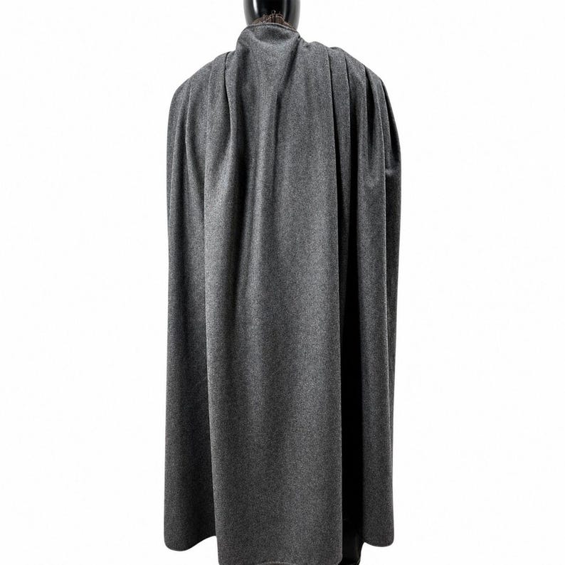 May include: A gray cape made of a soft, textured fabric. The cape is draped over a black mannequin, showcasing its full length and flowing design. The cape's color is a medium gray, and it appears to be made of a wool or similar material.