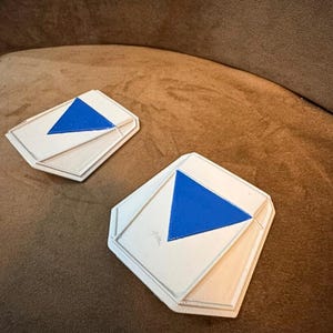 May include: Two white and blue geometric coasters with a blue triangle design. The coasters have a layered design and are set on a brown surface. The coasters are a decorative item.