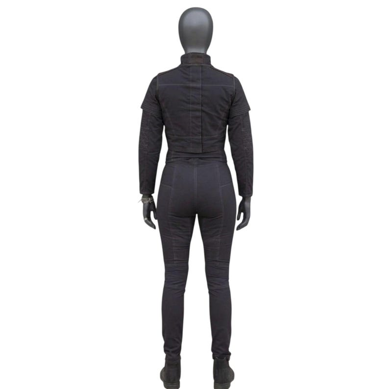 May include: A black jumpsuit displayed on a mannequin. The jumpsuit features a high collar, long sleeves, and fitted pants. The fabric appears to be textured, with visible stitching details. The mannequin is wearing black gloves and boots.