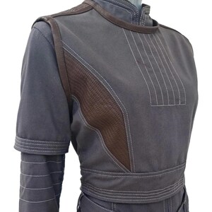 K&ouml;nnte beinhalten: Ein graues zweiteiliges Outfit mit braunen Lederakzenten. Das Oberteil hat einen hohen Kragen und kurze &Auml;rmel mit wei&szlig;en Steppdetails. Das Outfit umfasst eine taillierte Jacke und eine passende Hose, die ein ma&szlig;geschneidertes Design zeigen.