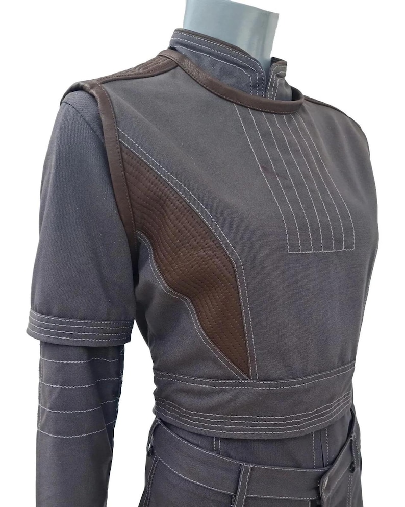 K&ouml;nnte beinhalten: Ein graues zweiteiliges Outfit mit braunen Lederakzenten. Das Oberteil hat einen hohen Kragen und kurze &Auml;rmel mit wei&szlig;en Steppdetails. Das Outfit umfasst eine taillierte Jacke und eine passende Hose, die ein ma&szlig;geschneidertes Design zeigen.