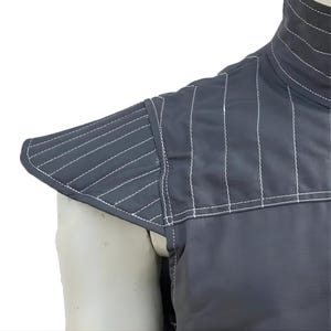 May include: Close-up of a dark gray garment with white stitching. The garment features a high collar and padded shoulder detail. The stitching creates a quilted effect, adding texture to the design. The fabric appears smooth and durable.
