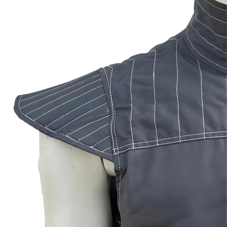 May include: Close-up of a dark gray garment with white stitching. The garment features a high collar and padded shoulder detail. The stitching creates a quilted effect, adding texture to the design. The fabric appears smooth and durable.