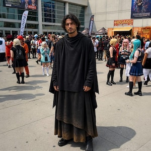 May include: A person wearing a black hooded cloak and layered skirt, standing in a crowd. The outfit appears to be made of a heavy fabric. The person is wearing black boots. The background shows a convention center with people in costumes.