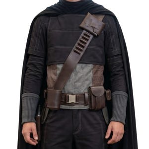 May include: A black costume featuring a cape, long-sleeved shirt, and pants. The outfit includes a brown leather shoulder strap with bullet holders, a belt with a silver buckle, and various pouches. Gray knitted wrist cuffs complete the look.