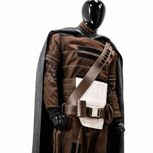 May include: A brown costume with a gray cape, displayed on a mannequin. The costume features a brown jumpsuit with black accents, a white padded chest piece, and a brown leather belt and shoulder strap. The sleeves have geometric stitching.