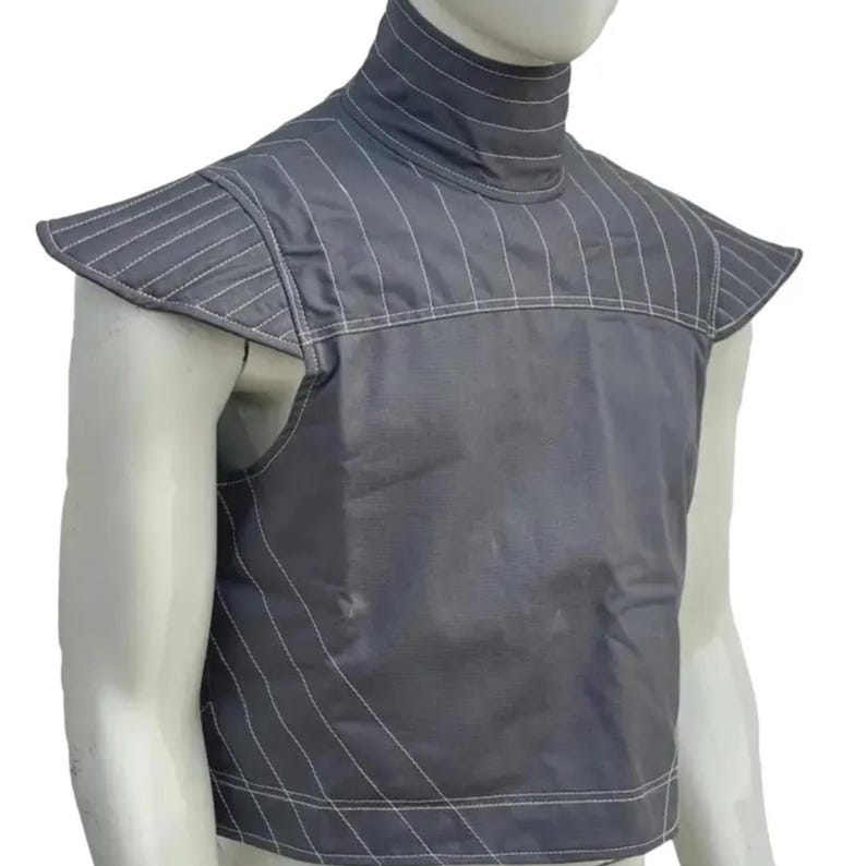 May include: A gray vest with a high collar and shoulder pads. The vest is made of a smooth fabric and features vertical white stitching details. The shoulder pads have a unique, angular design. The vest is displayed on a mannequin.