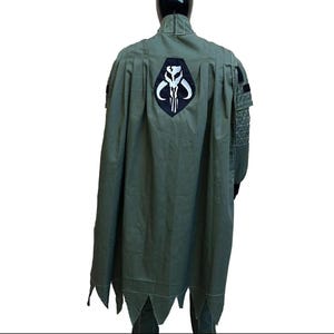 May include: Olive green cape with a black and white emblem on the back. The cape has a pointed hemline and is part of a costume. The costume is displayed on a mannequin.