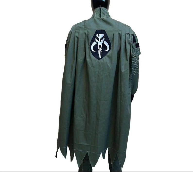 May include: Olive green cape with a black and white emblem on the back. The cape has a pointed hemline and is part of a costume. The costume is displayed on a mannequin.