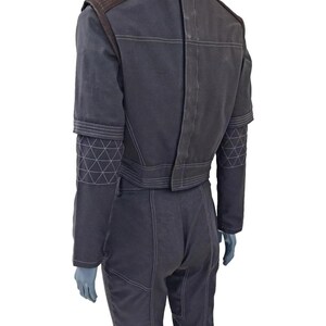 K&ouml;nnte beinhalten: Dunkelgrauer Overall mit wei&szlig;en Steppdetails. Die Jacke hat braune Schulterpatches und geometrische Muster an den &Auml;rmeln. Die Hose hat Taschen und sichtbare N&auml;hte. Das Outfit wird auf einer Schaufensterpuppe pr&auml;sentiert.