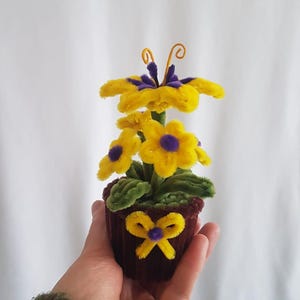 Può includere: Una composizione floreale fatta a mano in un piccolo vaso marrone. I fiori sono gialli con centri viola e le foglie sono verdi. Un fiocco giallo con un centro viola adorna il vaso. La composizione è realizzata con scovolini.