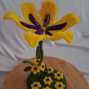 May include: A handcrafted, decorative flower sculpture with a large yellow and purple bloom. The flower sits atop a green stem with smaller yellow flowers and a green base. The sculpture is displayed on a wooden round base.