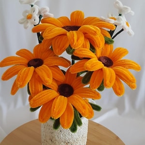 Può includere: Un bouquet di girasoli arancioni e marroni fatti a mano in un vaso bianco strutturato. I fiori hanno steli verdi e sono disposti su una superficie di legno. Sono inclusi anche piccoli fiori bianchi con centri gialli.