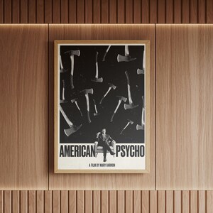 K&ouml;nnte beinhalten: Ein gerahmtes Poster mit dem Titel "AMERICAN PSYCHO" und einem Schwarz-Wei&szlig;-Bild von &Auml;xten und einem Mann im Anzug. Das Poster h&auml;ngt an einer Holzwand.