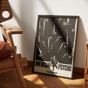 K&ouml;nnte beinhalten: Gerahmtes Poster mit dem Filmtitel "AMERICAN PSYCHO", das ein schwarz-wei&szlig;es Axt-Design und eine sitzende Figur zeigt. Das Poster befindet sich in einem schwarzen Rahmen und lehnt an einer wei&szlig;en Wand neben einem Holzstuhl.