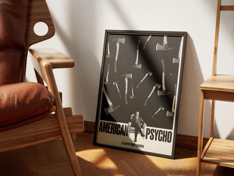 K&ouml;nnte beinhalten: Gerahmtes Poster mit dem Filmtitel "AMERICAN PSYCHO", das ein schwarz-wei&szlig;es Axt-Design und eine sitzende Figur zeigt. Das Poster befindet sich in einem schwarzen Rahmen und lehnt an einer wei&szlig;en Wand neben einem Holzstuhl.