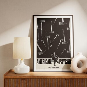 K&ouml;nnte beinhalten: Gerahmtes Poster mit dem Text "AMERICAN PSYCHO" und einem schwarz-wei&szlig;en Axtmuster. Ein Mann im Anzug sitzt in der Mitte. Eine wei&szlig;e Lampe und Vase stehen auf einem Holzschrank.
