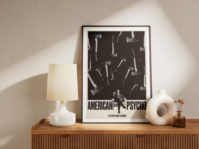 K&ouml;nnte beinhalten: Gerahmtes Poster mit dem Text "AMERICAN PSYCHO" und einem schwarz-wei&szlig;en Axtmuster. Ein Mann im Anzug sitzt in der Mitte. Eine wei&szlig;e Lampe und Vase stehen auf einem Holzschrank.