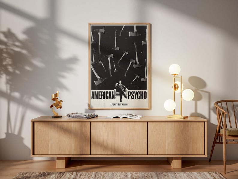 K&ouml;nnte beinhalten: Ein gerahmtes Filmplakat von "American Psycho" mit einem Schwarz-Wei&szlig;-Design mit &Auml;xten und einem Mann. Das Poster h&auml;ngt &uuml;ber einem hellen Holzschrank mit drei T&uuml;ren. Eine goldene Lampe mit kugelf&ouml;rmigen Gl&uuml;hbirnen und eine dekorative Skulptur stehen auf dem Schrank.