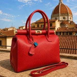 Peut inclure: Un sac à main en cuir rouge texturé doté de deux anses arrondies, d'une fermeture supérieure en métal doré et d'un petit cadenas bleu suspendu à une lanière en cuir. Une bandoulière rouge détachable est posée devant le sac.