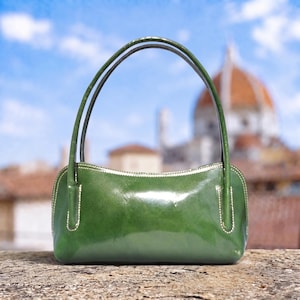 May include: A glossy, forest green handbag with a structured, rectangular body and rounded top corners. It features two thin, matching green shoulder straps attached to the front and back panels with visible, light-colored contrast stitching.