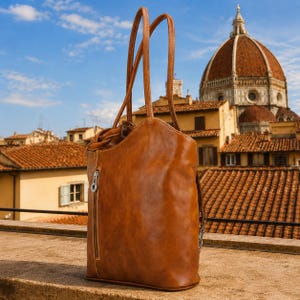 May include: A tan leather backpack with two long shoulder straps and a vertical zippered pocket on the front. The bag features visible stitching along the seams and a silver-toned metal zipper pull, showcasing a smooth, polished surface texture.