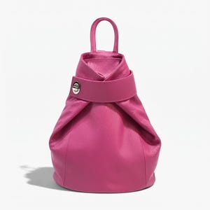 May include: A vibrant fuchsia pink leather backpack with a textured grain finish. The bag features a single top handle, a wide horizontal strap across the upper section, and a circular silver-toned metal turn-lock closure on the left side.