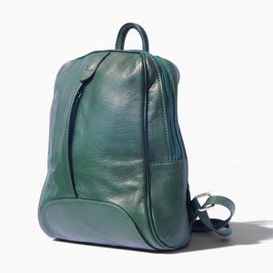 Puede incluir: Una mochila de cuero verde oscuro con textura lisa, asa superior, bolsillo frontal vertical con cremallera y correas ajustables con herrajes de metal plateado. La bolsa presenta una silueta redondeada y un compartimento principal.