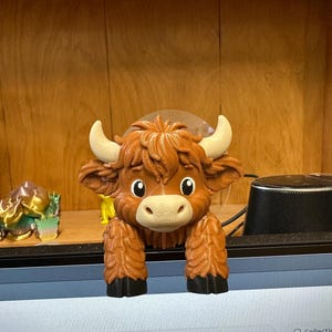 May include: A brown and tan Highland cow figurine with a friendly expression. The figurine has large, expressive eyes, small horns, and a fluffy coat. The cow is perched on a surface, with its front legs hanging down. The figurine is likely a decorative item or toy.