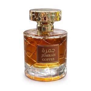 May include: A perfume bottle with a gold-colored geometric cap and a clear, textured glass body. The bottle contains a brown liquid and has a rectangular label with Arabic and English text: "Jumrah Coffee."