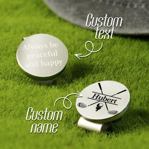 May include: Two silver golf ball markers on green grass. One marker has the text "Always be peaceful and happy." The other marker has the name "Hubert" and golf club graphics. The image also includes the words "Custom text" and "Custom name."
