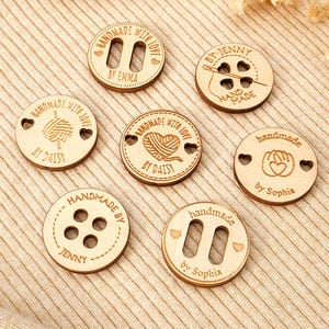 Könnte beinhalten: Eine Sammlung von Holzknöpfen mit verschiedenen Designs und Texten. Die runden Knöpfe tragen Aufschriften wie "Handmade with Love" und "Handmade by" sowie Namen. Einige Knöpfe haben Löcher, andere Schlitze, und einige zeigen Garn- oder Nähdesigns.