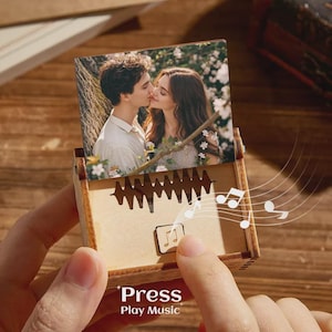 Könnte beinhalten: Eine Holz-Spieluhr mit einem Foto eines sich küssenden Paares. Die Box hat ein ausgeschnittenes Design und die Worte "Press Play Music" sind auf der Vorderseite aufgedruckt. Noten sind um die Box herum gezeichnet.