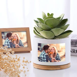 May include: A white ceramic planter with a wooden base, holding a green succulent plant. The planter features a printed photo of a couple. A wooden framed photo of the same couple is also present, along with dried flowers.