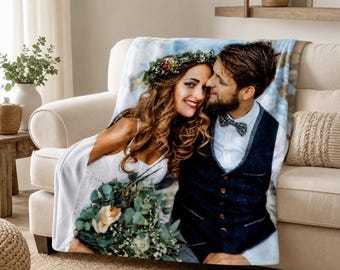 May include: A soft throw blanket featuring a printed photograph of a couple. The person on the left wears a white dress and a floral crown, holding a bouquet. The person on the right wears a white shirt, a dark blue vest, and a bow tie.
