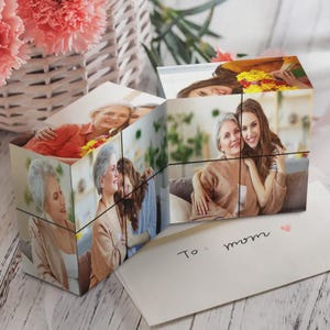 Peut inclure: Un cube photo personnalisé présentant plusieurs images imprimées de membres de la famille, dont une personne âgée et une plus jeune, posé sur une surface blanche à côté d'une enveloppe avec le texte manuscrit To: mom et un petit cœur rouge.