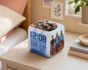 May include: A white cube-shaped digital alarm clock featuring custom photo prints on the top and side panels. The front display shows the time 12:08, the day TUE, the date 4/03, and a temperature reading of 26.1 degrees Celsius in blue text.