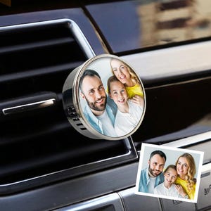 Op de afbeelding: Een ronde auto-luchtverfrisser met clip, voorzien van een gepersonaliseerde foto van een gezin van drie. De zilverkleurige metalen behuizing heeft zijdelingse ventilatieopeningen en is bevestigd aan een zwart ventilatierooster.