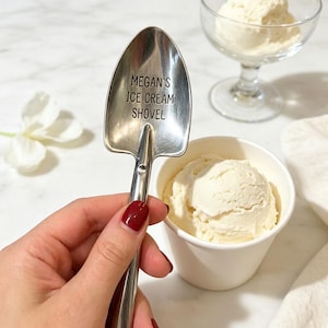 May include: A silver-toned metal spoon shaped like a miniature shovel with the text MEGAN'S ICE CREAM SHOVEL engraved on the bowl. The spoon is positioned next to a white paper cup filled with a scoop of vanilla ice cream and a glass dessert dish.