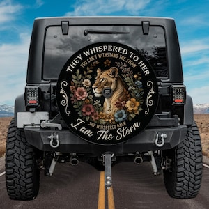May include: A black circular spare tire cover for a vehicle featuring a central illustration of a lioness surrounded by colorful flowers. The text reads: THEY WHISPERED TO HER YOU CAN'T WITHSTAND THE STORM I AM THE STORM SHE WHISPERED BACK.