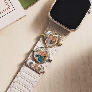 K&ouml;nnte beinhalten: Ein helles Smartwatch-Armband mit drei metallischen Anh&auml;ngern, die am Band befestigt sind. Jeder Anh&auml;nger zeigt ein anderes Foto: ein herzf&ouml;rmiger goldener Anh&auml;nger, ein runder silberner Anh&auml;nger und ein rechteckiger ros&eacute;goldener Anh&auml;nger mit Portr&auml;ts.
