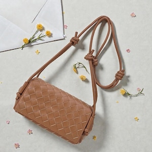 Puede incluir: Un bolso bandolera de cuero tejido de color marr&oacute;n con una correa anudada. El bolso rectangular presenta un patr&oacute;n cuadrado repetitivo. La correa tambi&eacute;n es marr&oacute;n y est&aacute; anudada en la parte superior y los lados. Flores amarillas y rosas est&aacute;n esparcidas alrededor del bolso.