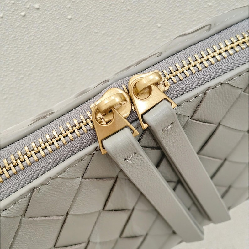 Pu&ograve; includere: Primo piano di un portafoglio in pelle grigia con un design intrecciato. Il portafoglio presenta una cerniera color oro e due tiralampo abbinati. La pelle ha una lucentezza sottile e l'estetica generale &egrave; minimalista ed elegante.