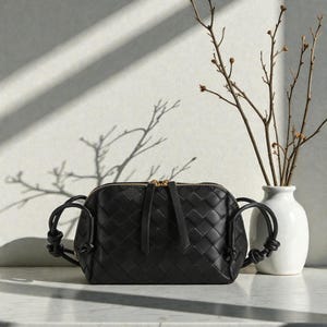 Pu&ograve; includere: Borsa a tracolla in pelle nera con design intrecciato. La borsa ha una cerniera dorata e manici annodati. Un vaso bianco con rami &egrave; sullo sfondo. La borsa &egrave; su una superficie bianca.