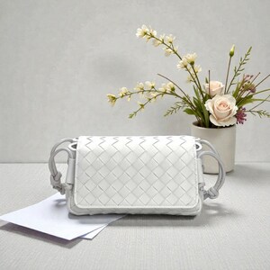 Puede incluir: Un bolso cruzado blanco de cuero tejido con cierre de solapa y correa gris. El bolso es rectangular y presenta un patr&oacute;n de tejido. Un jarr&oacute;n con flores est&aacute; en el fondo.