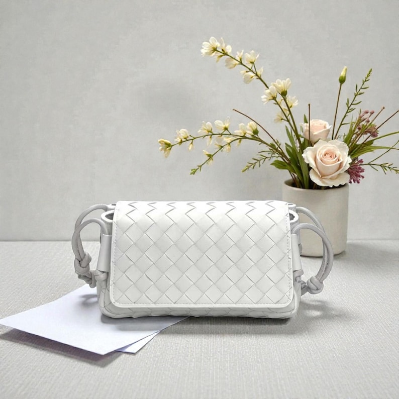 Puede incluir: Un bolso cruzado blanco de cuero tejido con cierre de solapa y correa gris. El bolso es rectangular y presenta un patr&oacute;n de tejido. Un jarr&oacute;n con flores est&aacute; en el fondo.
