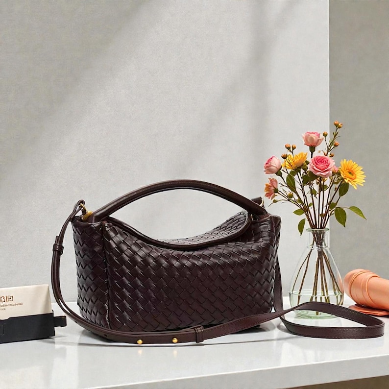 May include: A dark burgundy leather handbag with a woven pattern. The bag features a top handle and a long, adjustable shoulder strap. Gold studs are visible on the strap. A bouquet of flowers in a glass vase is in the background.