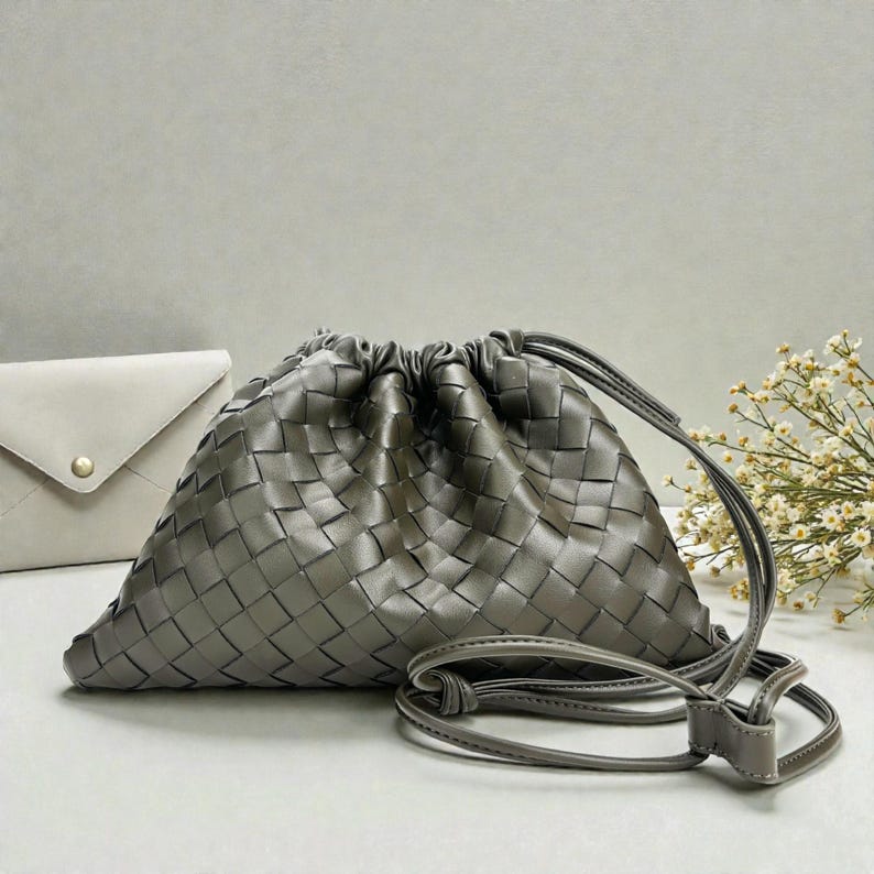 Puede incluir: Un bolso gris de cuero tejido con cord&oacute;n y correa larga para el hombro. El bolso tiene forma triangular y la parte superior fruncida. Un sobre peque&ntilde;o de color gris claro es visible en el fondo.