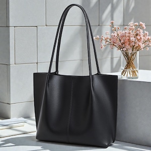 May include: A black tote bag with long handles. The bag is made of a smooth material and has a vertical seam down the center. A vase of pink flowers sits to the right of the bag. The background is a white tiled wall.