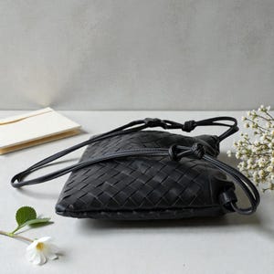 Puede incluir: Un bolso bandolera negro de cuero tejido con cierre de cord&oacute;n. El bolso tiene una correa larga y delgada y una forma rectangular. El bolso est&aacute; hecho de un material tejido y tiene una textura suave y flexible.